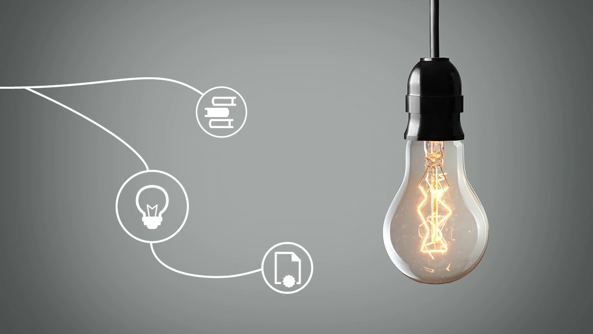 Una lampadina accesa è appesa a un cavo davanti a una parete grigia.